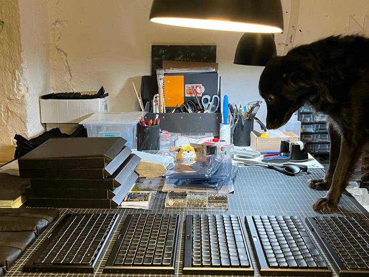 work table with DIY keyboards, random tools an boxes, dog standing with front paws on that table and looking exhausted